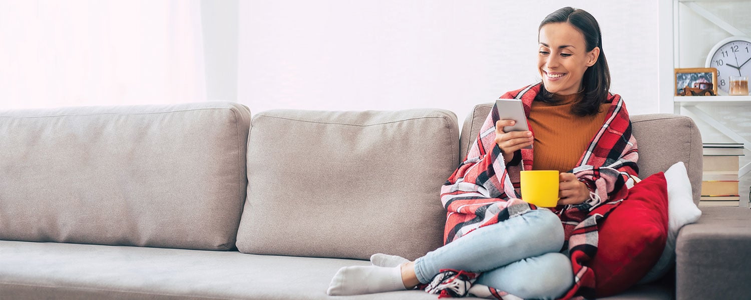 Woman sitting on couch, smiling at her phone. She's wearing a red plaid blanket